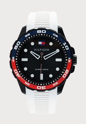 Ρολόι Tommy Hilfiger με μαύρο καντράν, λευκούς δείκτες ώρας, κόκκινο και μπλε στεφάνι, λευκό λουράκι από σιλικόνη και αντοχή στο νερό έως 100 μέτρα.