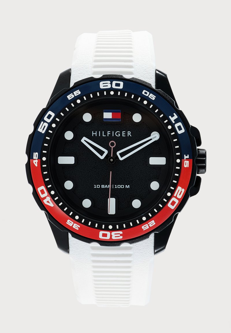 Ρολόι Tommy Hilfiger με μαύρο καντράν, λευκούς δείκτες ώρας, κόκκινο και μπλε στεφάνι, λευκό λουράκι από σιλικόνη και αντοχή στο νερό έως 100 μέτρα.