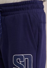 Temno modri trenirki, izdelani iz mehkega materiala; imajo elastiko v pasu, stranske žepke in bel tiskani logotip na stegnu.
