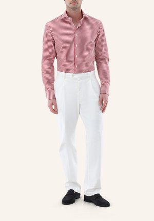 Mann trägt rotes und weißes, vertikal gestreiftes Hemd mit Knöpfen, weiße Hose und schwarze Loafers, steht vor einfachem Hintergrund.