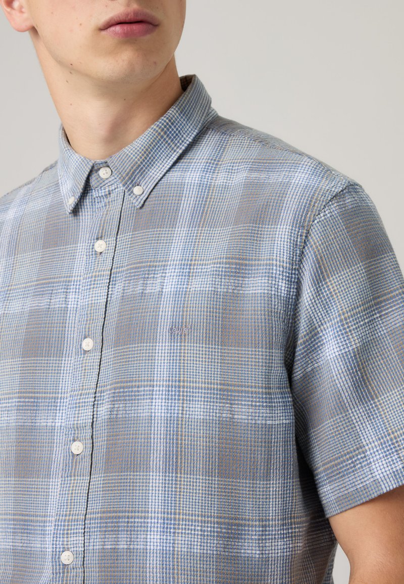 Homme portant une chemise à manches courtes boutonnée avec un motif à carreaux bleu, gris et blanc, col boutonné, fond neutre.