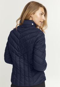 Námornícka modrá padded bunda s vysokým golierom, chevron prešívaním a detailom na cvoky na krku. Hladká textúra.