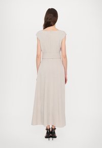 Ljusbeige klänning med kort ärm och figurnära liv, bälte i midjan som flödar ner till golvet, textilier med struktur, minimalistisk design. Svarta klackskor.