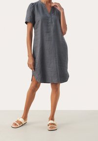 Robe en lin gris avec un col en V et des manches courtes bouffantes, dotée d'une fente latérale pour faciliter le mouvement. Portée avec des sandales plates de couleur claire.