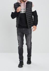 Chaleco acolchado negro sobre una sudadera negra con un acento rosa. Combinado con jeans oscuros y zapatillas deportivas negras. Diseño simple y moderno.