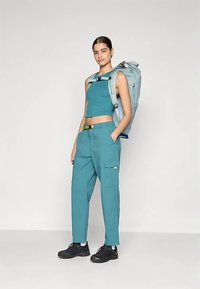 Γυναίκα που στέκεται, φοράει τιρκουάζ αμάνικο μπλούζα και παντελόνι cargo με μαύρες ορειβατικές μπότες, κρατώντας ένα ανοιχτό μπλε σακίδιο για πεζοπορία.