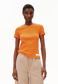 Oranje T-shirt met korte mouwen, met een geribbelde textuur, ronde halslijn en een wit label op de zoom. Gepaard met beige joggingbroeken met trekkoord.