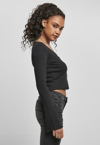 Musta ribbed pitkähihainen crop-top, jossa on aaltoileva helma ja levenevät hihat, yhdistettynä tummiin farkkuihin harmaata taustaa vasten.
