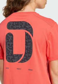 Koralowy t-shirt z krótkim rękawem z dużym czarnym graficznym logo i napisem "2026 TEAM D ADIDAS" na plecach. Materiał bawełniany, gładka faktura.