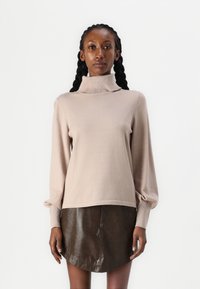 Beige garbónyakú pulóver hosszú, puffos ujjaival és bordázott mandzsettákkal; egy barna bőr mini szoknyával viselve, amely ívelt szegéllyel rendelkezik.