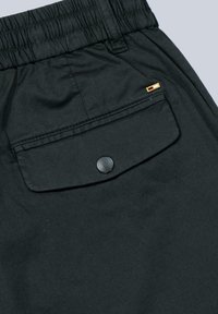 Black shorts featuring an elastic waistband, a rear flap pocket with a black snap button, and a small gold accent detail. Smooth texture.
