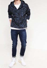 Veste à capuche légère, bleu marine, avec un motif texturé, poche avant et capuche ajustable, associée à un jean foncé et des chaussures blanches.