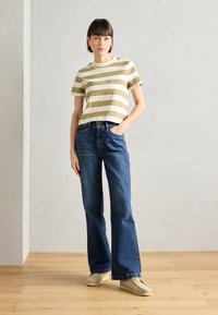 T-shirt a righe olive e crema abbinato a jeans in denim blu a zampa d'elefante. Il modello sta in piedi con le mani in tasca, indossando scarpe di colore chiaro.