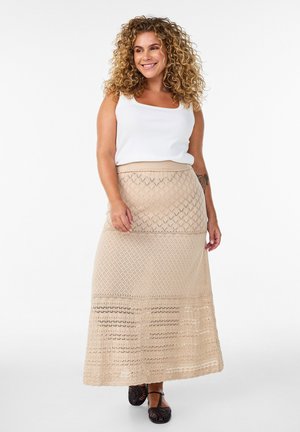 Kiharahiuksinen nainen hymyilee, pukeutuneena valkoiseen hihattomaan toppiin, beigen tekstuuriin ja mustiin kudottuihin suljettuin varvassandaaleihin, seisoo valkoista taustaa vasten.