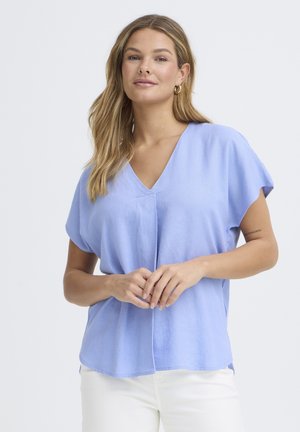 Žena so svetlohnedými vlasmi, ktorá má na sebe zlaté kruhové náušnice, voľný svetlomodrý top s výstrihom do V a biele nohavice, stojaca pred jednoduchým pozadím.