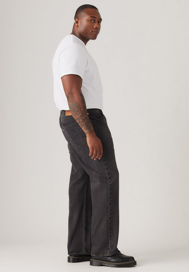 Jean en denim noir avec une coupe droite, porté avec un t-shirt blanc à manches courtes et des bottes noires. Tatouage visible sur le bras droit.