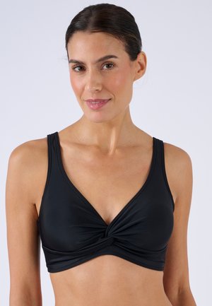 Mujer con un top corto negro con nudo frontal y tirantes anchos, sonriendo suavemente frente a un fondo claro y simple.