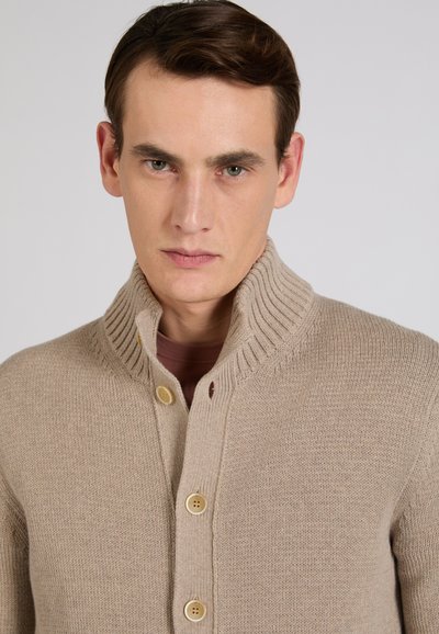 Giovane uomo con capelli castani corti che indossa un cardigan in maglia beige abbottonato con collo alto su uno sfondo semplice.