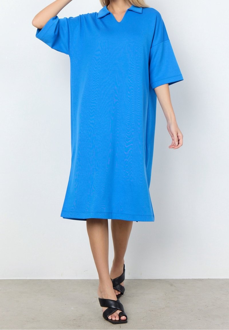 Personne portant une robe ample bleu vif mi-longue avec manches courtes et col, associée à des sandales noires à talons ouverts.