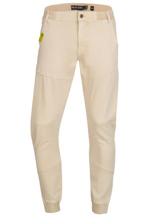 Beige tapered pants in a soft fabric, featuring stitched accents and elastic cuffs. Includes a front button closure and five pockets.