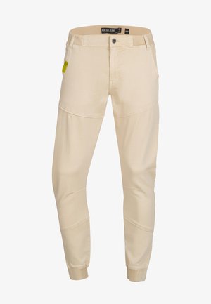 Pantalons beige fuselés en tissu doux, avec des accents cousus et des poignets élastiques. Comprend une fermeture à bouton à l'avant et cinq poches.