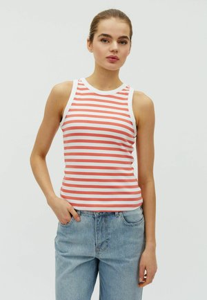 Ung kvinde med håret bundet tilbage iført en hvid ærmeløs tanktop med koralrosa striber og lyseblå jeans, stående med den ene hånd i lommen.