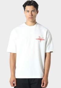 Camiseta blanca de manga corta con un diseño gráfico rojo y el texto "PAS DE MONACO" en el pecho. Fabricada en una suave tela de algodón.