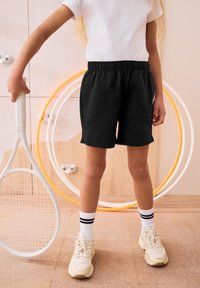 Shorts noirs en mélange de coton avec une taille élastique, des poches latérales et une coupe décontractée, associés à un t-shirt blanc et des baskets beiges.