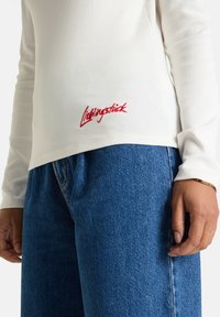 Person, der ein weißes geripptes Langarmoberteil mit rober "Lightingstick"-Stickerei und blauen Jeans trägt, mit entspanntem Arm an der Seite.