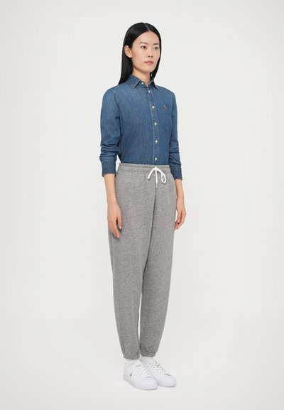 Džínová košeľa na gombíky, sivé tepláky s vrkočovým pásom a biele tenisky. Model stojí pred jednoduchým bielym pozadím.