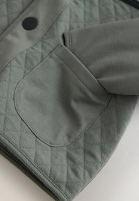 Quiltet grøn jakke med en glat, mat lomme detalje. Har en rund krave og struktureret stof, hvilket giver den et lagdelt udseende.