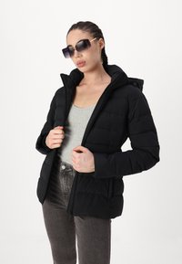 Manteau en duvet noir avec capuche, présentant une texture matelassée, une fermeture éclair et des poches latérales. Porté par-dessus un débardeur gris et associé à un jean gris.