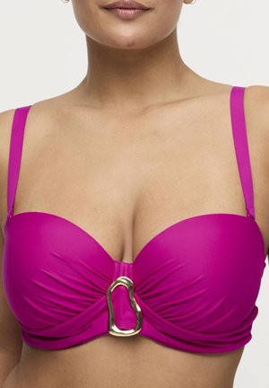 Vrouw draagt een felroze bikini top met geplooide stof en een groot onregelmatig gouden gespdetail aan de voorkant.