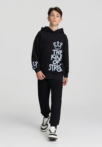 Czarna bluza z kapturem z białym napisem graficznym "KRÓL ULICY" i wzorem korony, w połączeniu z czarnymi spodniami i czarno-białymi sneakersami.