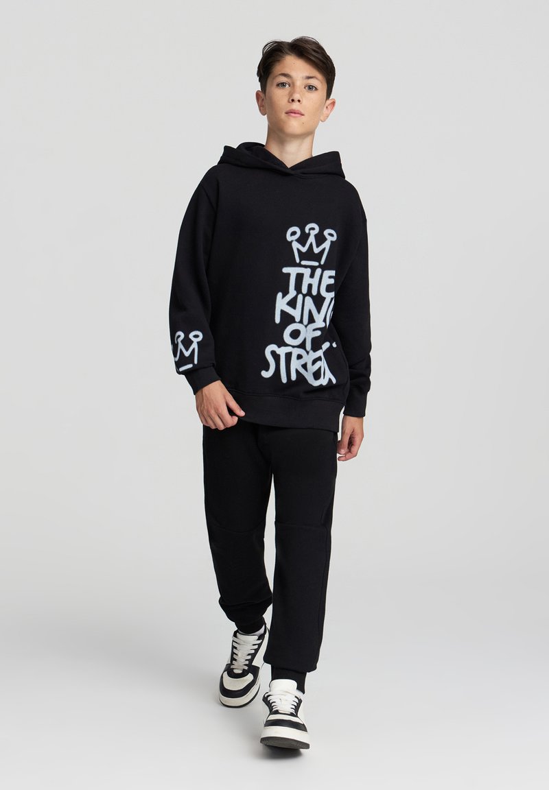 Czarna bluza z kapturem z białym napisem graficznym "KRÓL ULICY" i wzorem korony, w połączeniu z czarnymi spodniami i czarno-białymi sneakersami.