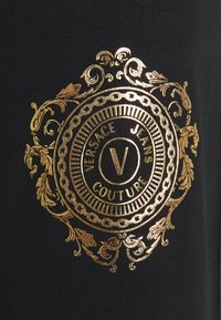 Goldgeprägtes Logo auf schwarzem Stoff mit filigranen Blumenakzenten, rundem Design und dem deutlich sichtbaren Schriftzug "VERSACE JEANS COUTURE".