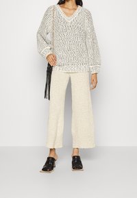 Strickpullover in Weiß und Schwarz mit V-Ausschnitt und langen Ärmeln. Strukturiert, lockere Passform. Cremefarbene weitgeschnittene Hosen in Knöchellänge. Schwarze Slipper.