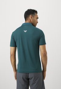 Vyras su trumpais tamsiais plaukais, vilkintis smaragdinio spalvos polo marškinėlius su baltu logotipu ant nugaros ir pilkas šortus, atsukęs nugarą.