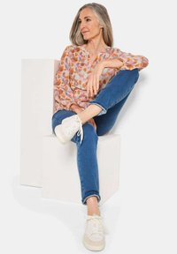 Bluse mit floralem Muster in sanften Rosa- und Orangetönen, kombiniert mit engen blauen Jeans und weißen Sneakers, sitzend auf einem weißen Würfel.