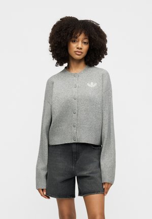 Frau mit lockigem Haar, die eine hellgraue, geknöpfte Strickjacke mit Logo und hoch taillierte dunkelgraue Jeansshorts trägt.