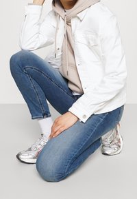 Persona de rodillas con sudadera beige claro debajo de una chaqueta blanca, jeans azules, calcetines blancos y zapatillas plateadas sobre un suelo gris claro.