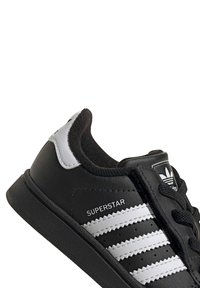 Čierne syntetické kožené tenisky s bielymi akcentmi, ktoré majú tri pruhy, textúrovanú gumovú podrážku a nápis 'SUPERSTAR'.