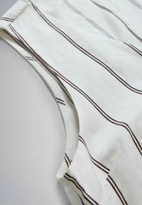Tissu blanc à fines rayures verticales marron, montrant un gros plan de la couture de l'épaule et de l'emmanchure d'un vêtement sans manches.