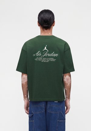 Vitiligosta kärsiv henkilö, jolla on yllään tummanvihreä Air Jordan -t-paita ja siniset farkut, selin yksiväristä taustaa vasten.