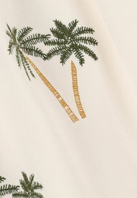 Tissu crème avec des palmiers brodés verts et des accents de troncs bruns, mettant en valeur des coutures détaillées et une texture lisse.