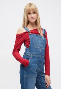 Spijkerbroekoveralls met verstelbare bandjes, voorzak en een aansluitend rood shirt met lange mouwen eronder. Lichtblauwe kleur en ontspannen pasvorm.