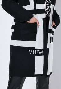 Lång svart och vit stickad cardigan med geometriskt mönster, med en ficka och ordet "VIEW" broderat i vitt på framsidan.