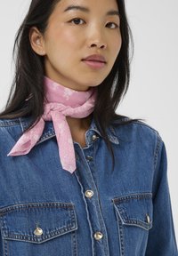 Sciarpa rosa a fantasia annodata attorno al collo, indossata su una camicia di jeans blu con dettagli in bottoni argentati e tasche sul petto.