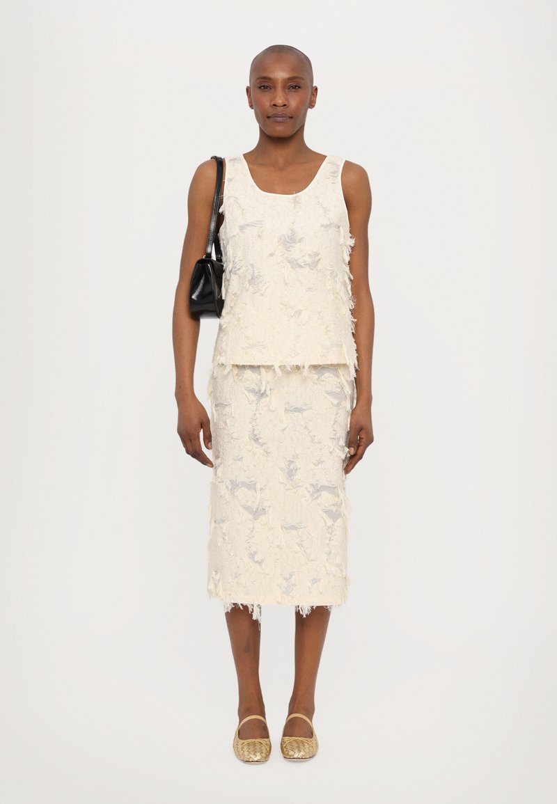 Crème textuur tweedelige outfit met een mouwloze top en een rok tot op de knie met rafelige randen. Draagt het met een zwarte tas en gouden schoenen.