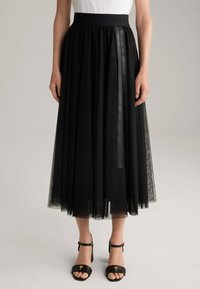 Jupe midi en tulle noir avec une taille élastique, présentant un logo et un détail en ruban satiné. Portée avec des sandales à talons noires.